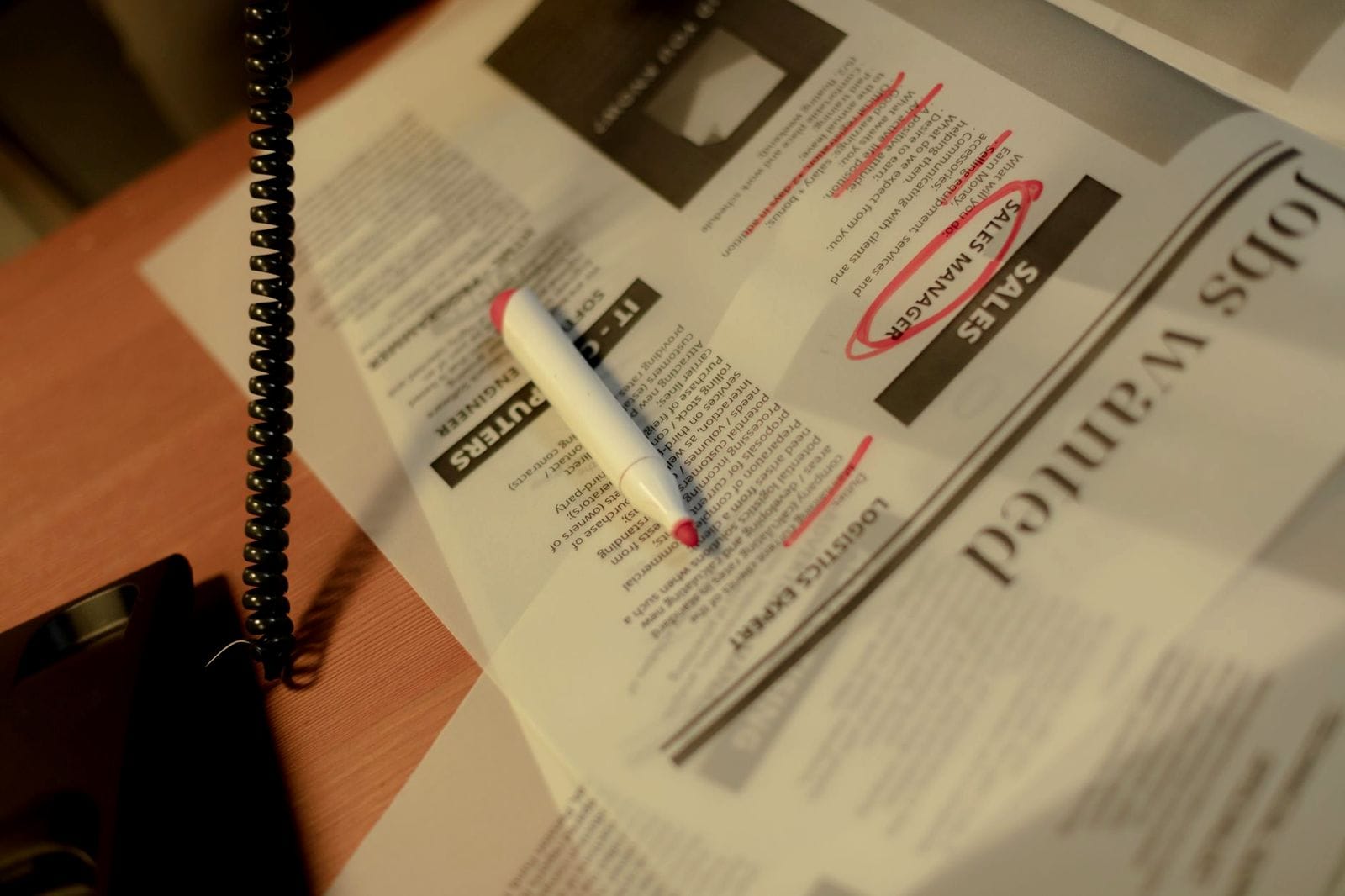 A newspaper jobs page with a sales manager listing circled in red marker, a capped marker lying across the listings and a coiled phone cord beside them