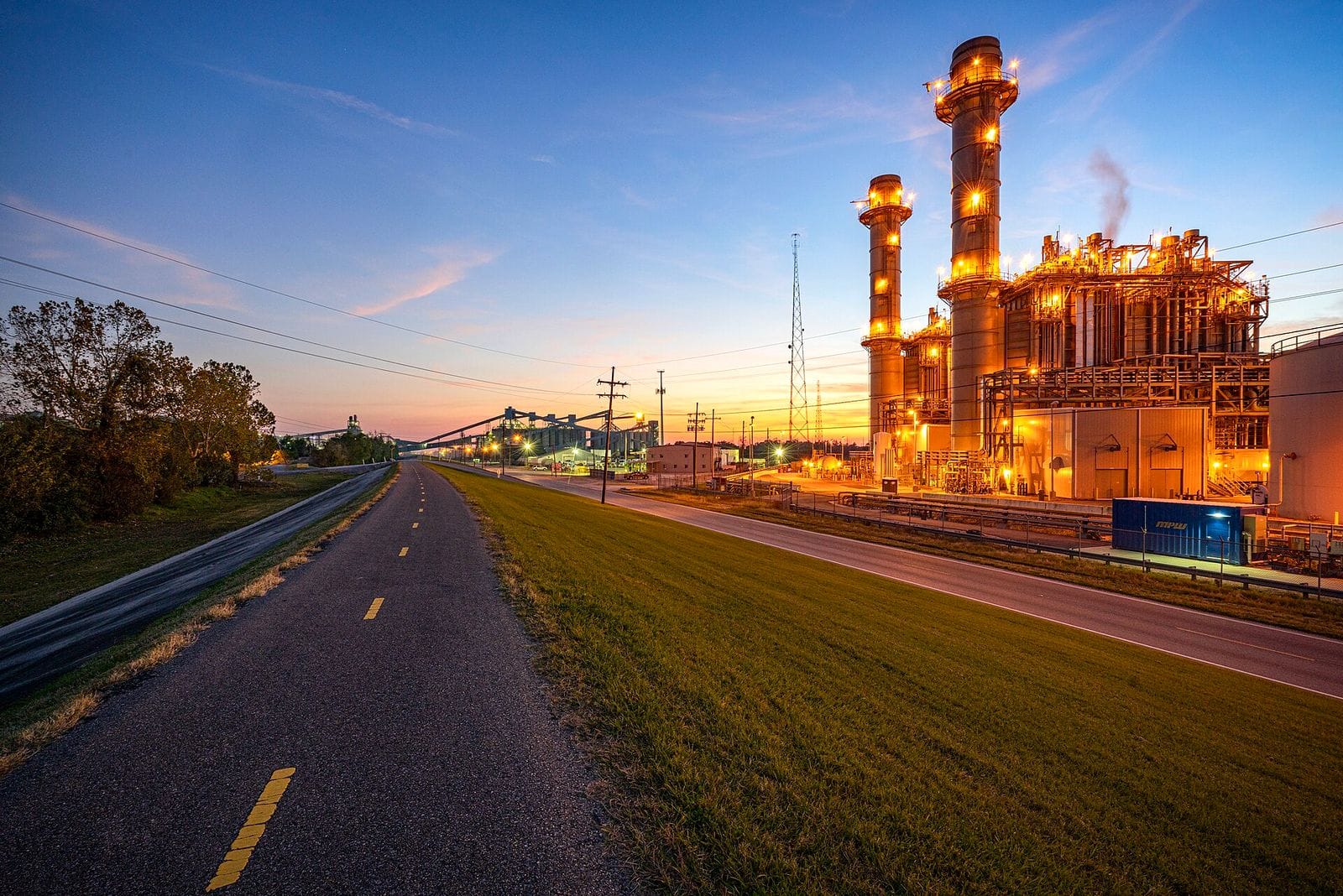 Gas-fired power plant with two tall exhaust stacks lit up at dusk, seen from a levee-top road with grass on either side