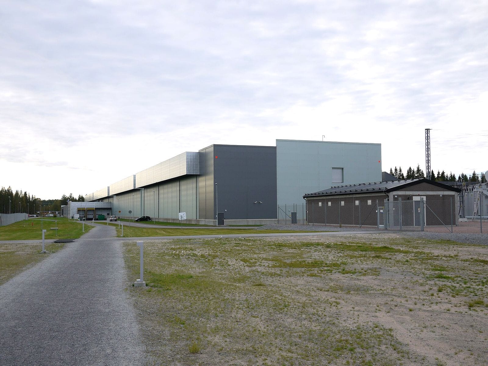Long, windowless grey data center building behind a gravel path and patchy grass under an overcast sky, with a smaller brown outbuilding and a lattice mast beside it