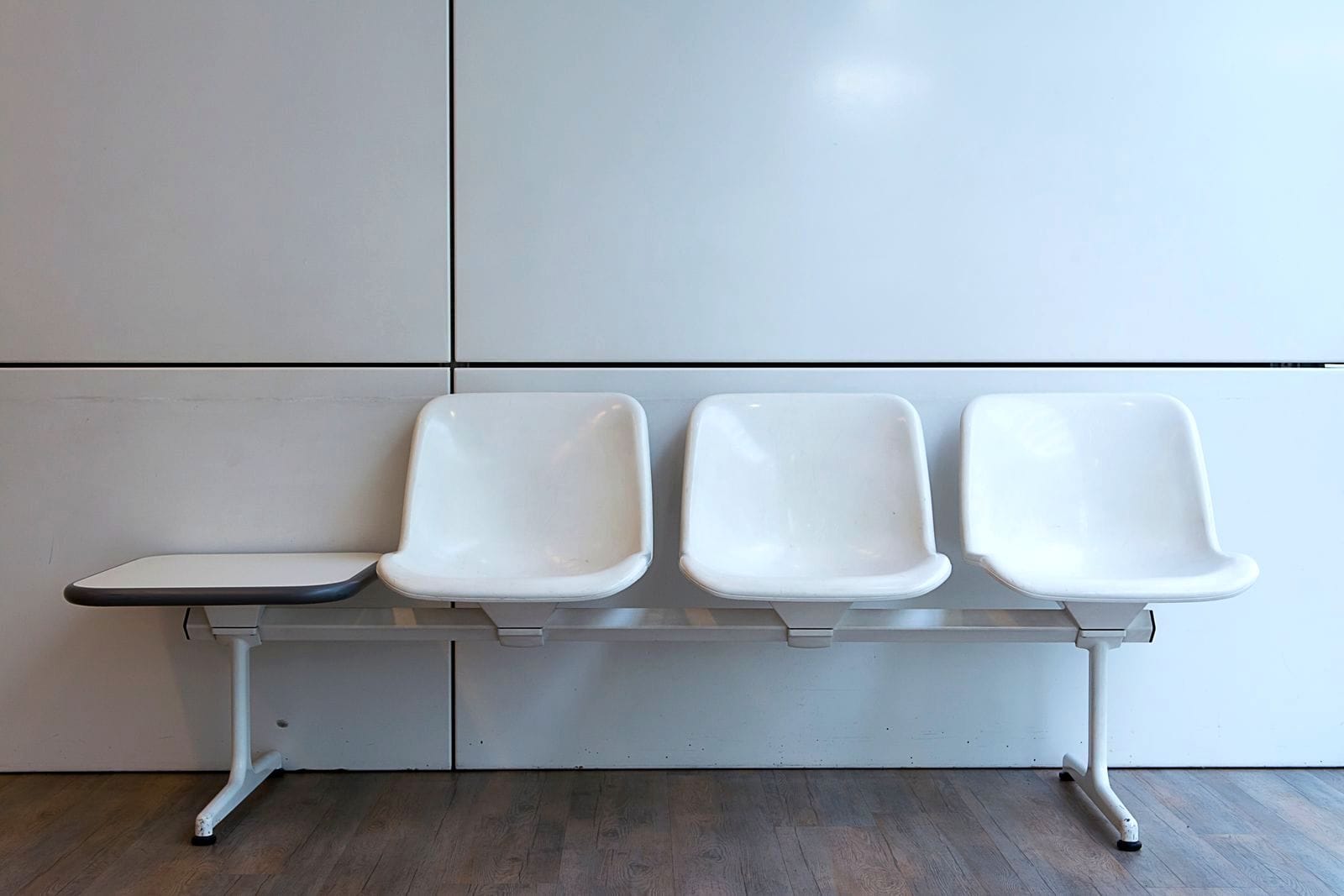 Row of three empty white plastic waiting-room chairs with an attached side table against a white wall